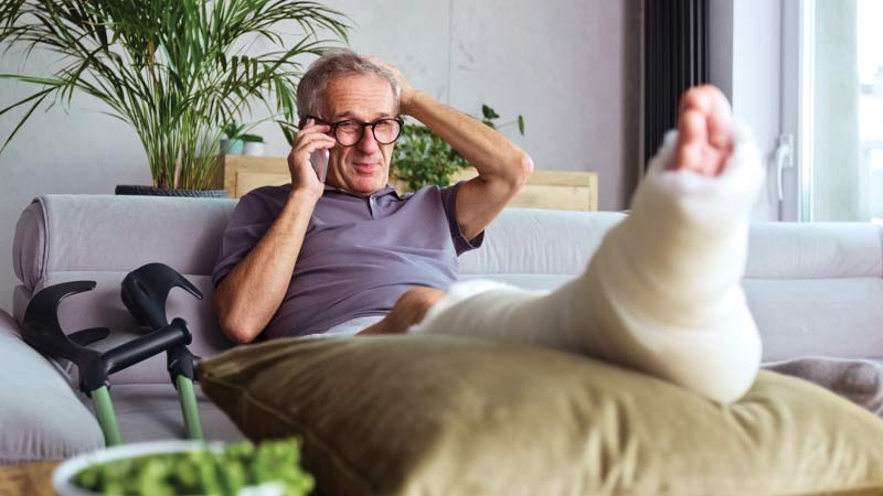 Homem senior sentado num sofá ao telemóvel, com a perna engessada sobre uma almofada; muletas ao lado